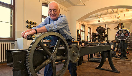 A man turns a large gear on a historical machine. It is very long, extending far back into the room. 