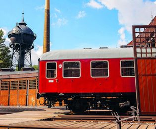 The gate to the Engine Shed, or roundhouse, is open, and a red compartment car is half inside. In the background, the Shed’s curved wall, a chimney, and a water tower can be seen.