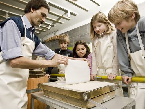 Vier Kinder schauen einem Vorführer dabei zu, wie er behutsam ein frisch geschöpftes Blatt Papier von einem Flies abhebt. Der Vorführer trägt eine Arbeitsschürze und hält das Papier vorsichtig mit den Händen an zwei Ecken fest.
