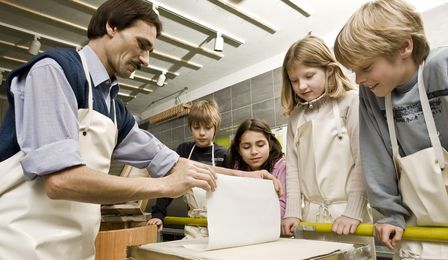 Vier Kinder schauen einem Vorführer dabei zu, wie er behutsam ein frisch geschöpftes Blatt Papier von einem Flies abhebt. Der Vorführer trägt eine Arbeitsschürze und hält das Papier vorsichtig mit den Händen an zwei Ecken fest.