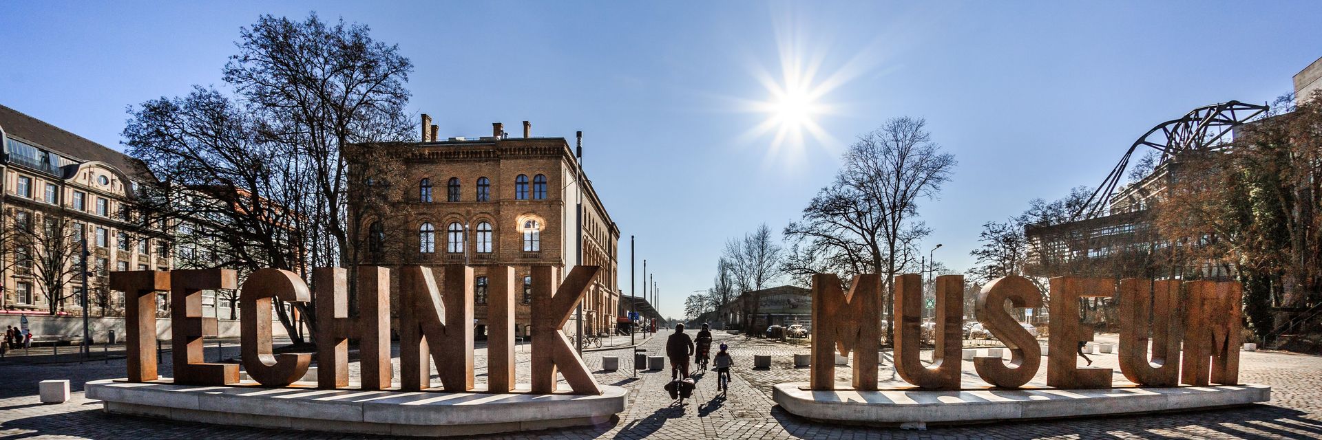 Auf einem gepflasterten Platz sieht man eine aus Metallbuchstaben erstellte Wortskulptur, die sich „Technik Museum“ liest. Im Hintergrund ein Backsteingebäude.