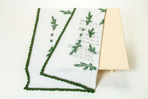 This rectangular tablecloth is made from white cotton. It is embroidered with green fir branches, which were applied by hand. An English language imprint on one part of the tablecloth is noteworthy. It shows that the tablecloth had previously been a flour sack. 