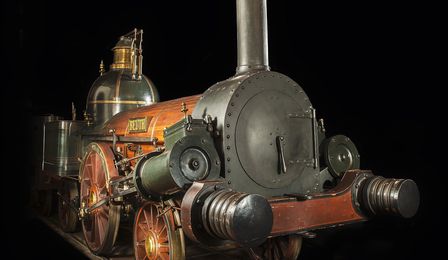Eine mächtige Dampflokomotive aus metallenen und hölzernen Bauteilen vor schwarzem Hintergrund