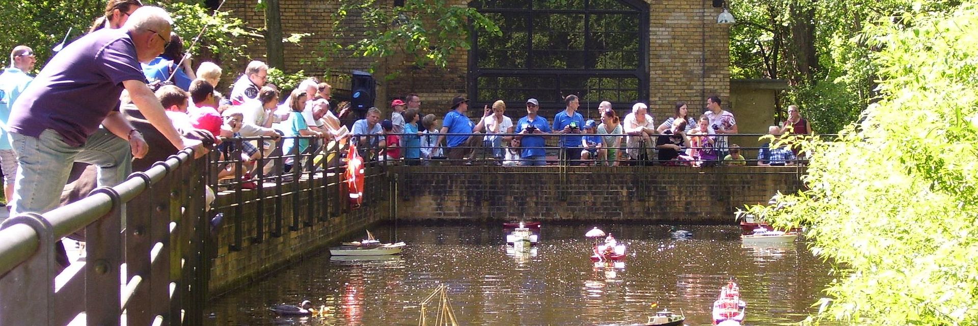 Besucherinnen und Besucher schauen auf den Museumteich im Park, auf dem kleine Modellschiffe hin und her fahren.