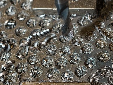 The metal drill has made numerous indentations in the surface of the tool table. The table is littered with metal shavings and small scraps left over from the last drilling.