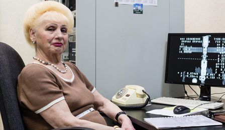 A heavily made-up elderly woman with short blond dyed hair is sitting at a desk on which there is an old telephone, a computer screen with numbers and a picture of the ISS.