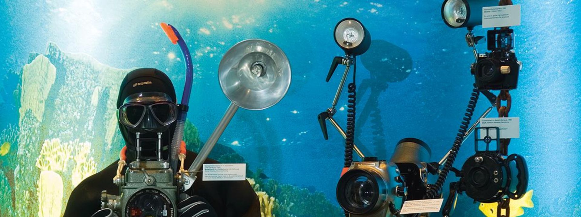 Blick in einen Schaukasten mit Unterwasseratmosphäre. Zu sehen ist ein lebensgroßer Taucher in schwarzem Taucheranzug und Taucherbrille mit Schnorchel und Unterwasserkamera in der Hand. Rechts daneben sind weitere Unterwasserkameras angebracht. 