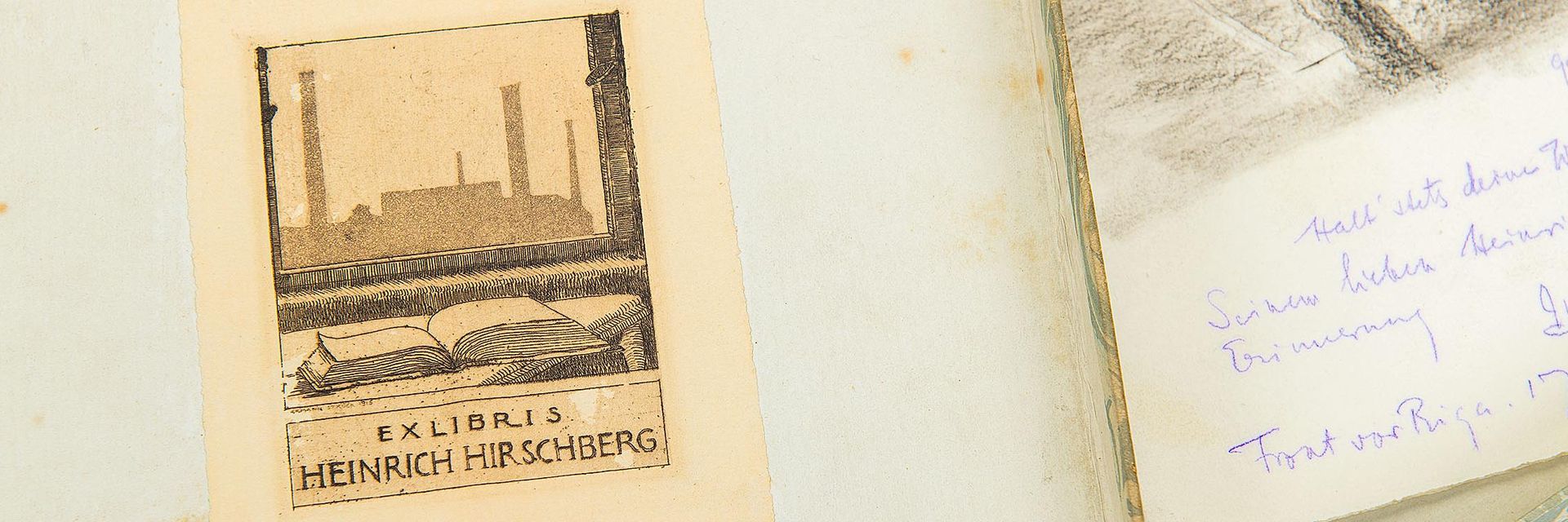 Aufgeklappte Schachtel, darin rechts eine Zeichnung mit einer Widmung. Links ist die Deckelinnenseite zu sehen, auf der Heinrich Hirschbergs Exlibris klebt. Es zeigt im Vordergrund ein aufgeschlagenes Buch auf einem Tisch, im Hintergrund ist durch einen Fensterrahmen eine Fabrik zu erkennen.