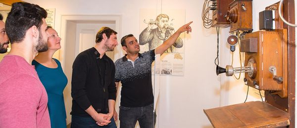 A group of museum visitors looks at various historical telephones hanging on a wall. One of the visitors has his arm outstretched and is pointing at a telephone.