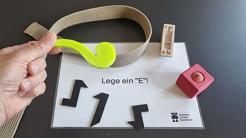 Hand mit grünem Werkzeug über einer Experimentierstation des Science Center Spectrum mit der Aufgabe „Lege ein ‚E‘!“ und mehreren geometrischen Formteilen.