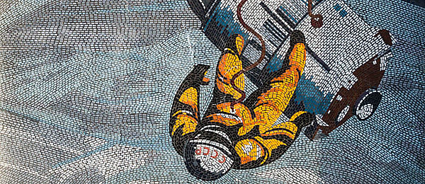 Das Mosaik zeigt einen Kosmonauten im gelben Anzug, der im All über der Erde schwebt. Über einen Schlauch ist er mit der dahinter schwebenden Raumkapsel verbunden.