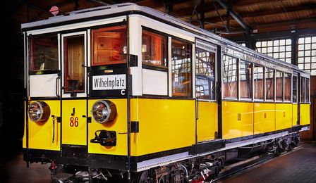 An historic subway car stands on a track in an engine shed. The square-shaped car has a yellow exterior and a white interior. 