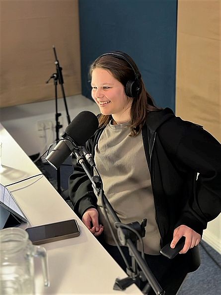 Eine junge Person mit braunen schulterlangen Haaren sitzt in einem Tonstudio vor einem Mikrofon und lächelt.