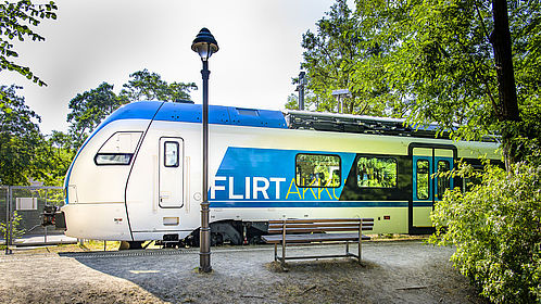 Seitenansicht von einem blau-weißen Zug mit der Aufschrift "FLIRT Akku". Der Zug steht auf einem Bahnsteig im Grünen ein, davor eine Laterne und eine Bank.