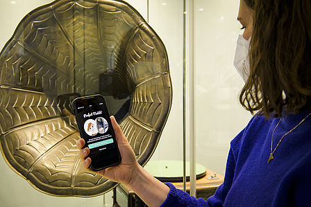 Eine Frau steht vor einem Grammofon, in der Hand hält sie ein Smartphone, auf dem Screen ist "Perfect Match" zu lesen.