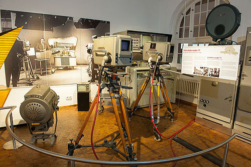 Zwei große Kameras und ein Scheinwerfer. An der Wand hängt eine schwarz-weiß Großaufnahme eines Fernsehstudios in den 1950er Jahren.