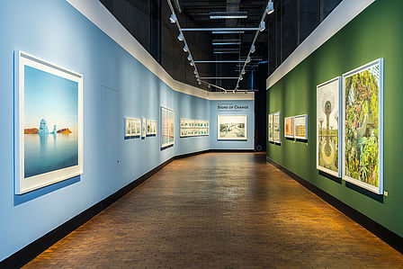 Blick in die Ausstellung "Signs of Change", links hängen Fotos an einer hellblauen Wand, rechts an einer dunkelgrünen Wand.