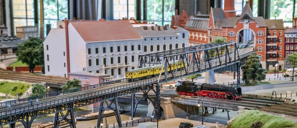 A detailed model train set. An elevated railway bridge with a yellow train on it crosses over railroad tracks, a canal, and a road. Model houses and miniature cars, trains, and boats populate the rest of the model.