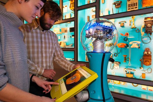 A woman and a man stand in front of a display case with Tim, the museum robot. The woman is using Tim’s touch screen, which shows an old, orange rotary phone.