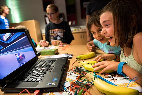 Kinder sitzen an einem Tisch vor einem Laptop. Auf dem Bildschirm ist ein Spiel mit einem Rennauto zu sehen. Die Kinder steuern das Spiel mit Bananen, die auf dem Tisch liegen und über Kabel und ein Gerät mit dem Laptop verbunden sind.