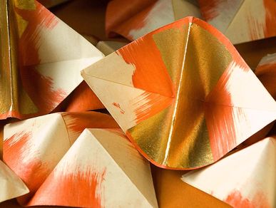 A pile of paper boats. From the pointed ends to the middle, the boats go from white to red. The central fold is embellished with gold.