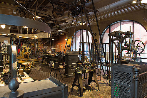 A view of the Historical Machine Shop, featuring several metalworking machines. Mounted to the ceiling are the line shaft and the various wheels and belts that transmit power from the steam engine to the machines.