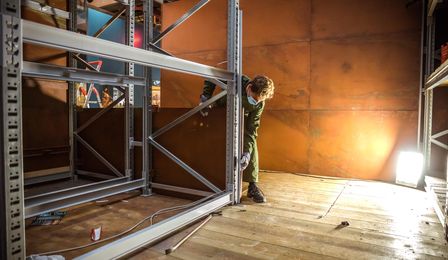 A man in green overalls is dismantling a metal shelf. A brown metal wall can be seen in the background.