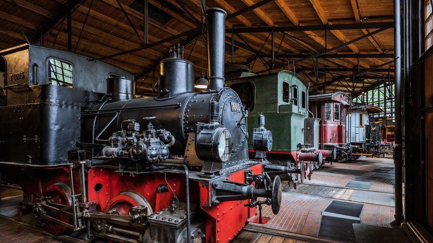 Blick in die Schienenverkehrs-Ausstellung: Eine historische schwarzrote Lokomotive, und mehrere Abteilwagen stehen auf Schienen nebeneinander aufgereiht.