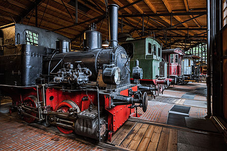 Blick in die Schienenverkehrs-Ausstellung: Eine historische schwarzrote Lokomotive, und mehrere Abteilwagen stehen auf Schienen nebeneinander aufgereiht.