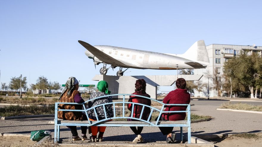 Four women are sitting on a park bench with their backs to the viewer. They are looking at a silver aircraft mounted on a concrete pedestal.