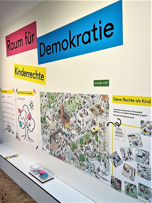 Blick in einen hellen Raum mit Plakaten an den Wänden. Darüber die Schriftzüge "Raum für Demokratie" und "Kinderrechte"