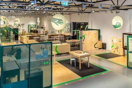 Blick in den Ausstellungsbereich "Reparieren!" mit seinen hellen Holzmöbeln, Werkzeugtischen und Grafik-Icons, die im Ausstellungsraum Orientierung bieten.