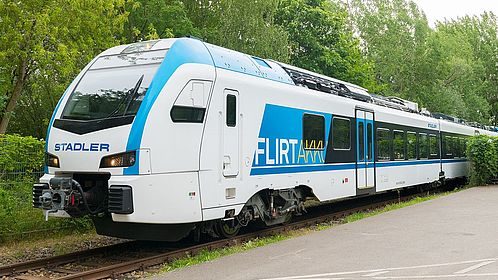 Dreiviertelansicht eines blau-weißen Zugs mit den Aufschriften „Stadler“ und „FLIRT Akku“. Der Zug fährt auf einem Gleis im Grünen, davor ist eine asphaltierte Fläche zu sehen.