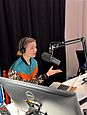 Moderatorin Jana Forkel sitzt in einem Tonstudio an einem Tisch mit einem Studiomikrofon und einem Laptop. Sie trägt eine bunt gemusterte Jacke und spricht in das Mikrofon.