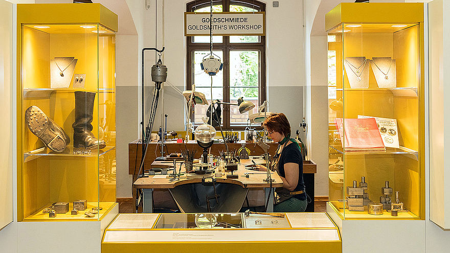 Between two yellow display cases containing a variety of objects, a table with numerous jewelry tools can be seen. There is a woman sitting at the table, working.