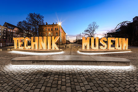 Auf einem gepflasterten Platz sieht man eine aus Metallbuchstaben erstellte Wortskulptur, die sich „Technik Museum“ liest. Im Hintergrund ein Backsteingebäude. Der Himmel ist durch die einsetzende Dämmerung dunkelblau. Die Wortskulptur ist beleuchtet.