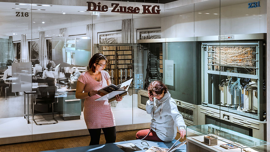 A view of the Computers exhibition. A woman sits on a ledge at an audio station, holding a handset to her ear. Another woman stands next to her, reading a large brochure. In the background, tall display cases containing historical computers can be seen. The words “Die Zuse KG” (The Zuse KG Company) hang from the ceiling.