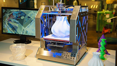 Ein 3D-Drucker aus silberglänzendem Metall. Darauf befindet sich eine weiße, gedruckte Vase.