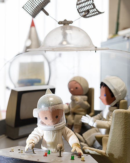 Das Sandmännchen im Kosmonautenanzug steht an einem Kontrollpult mit Schalthebeln. Im Hintergrund sitzen zweit weitere Holzpuppen in einer Raumstation, darüber ein Glasdach mit Satellitenschüsseln. 