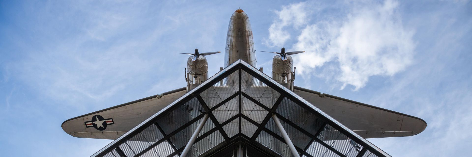 Blick von unten auf einen Neubau mit Glasfassade und Stahlstreben. Darüber hängt ein Flugzeug.
