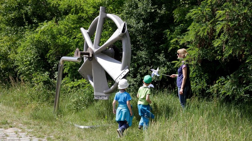 Two children and a woman look at a windmill hub. It is fastened to a metal support standing in high grass.