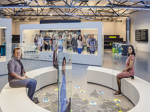 There is a circular bench in the middle of the CONNECT exhibit. Glass panels with life-size human figures on them are arranged around it. There is a graphic of a world map on the floor in the middle of the circle.