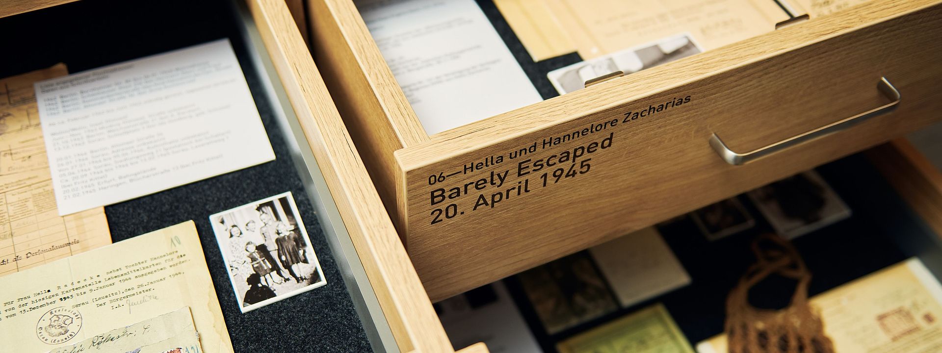 Blick in zwei geöffnete Schubladen mit Unterlagen, Briefen und Fotos. Auf einer Schublade ist zu lesen: Hella und Hannelore Zacharias. Barely Escaped, 20. April 1945