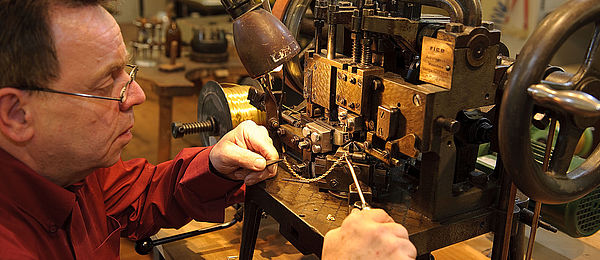 Ein Mann arbeitet an einer historischen Kettenmaschine. Mit zwei Pinzetten kontrolliert er die Glieder der entstehenden Kette. 