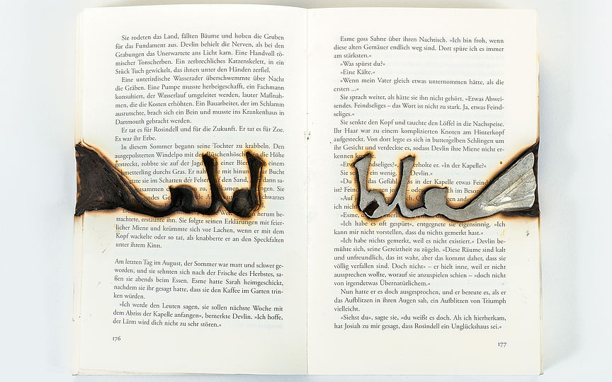 Ein Buch aus dessen Seiten das Wort „bla“ herausgeschnitten wurde. In den Holraum wurde Silber gegossen. Die Ränder sind von der Hitze verkohlt.