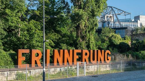 Umgeben von Bäumen ist eine Wortskulptur aus rostigem Metall zu sehen, deren Buchstaben das Wort "Erinnerung" bilden. Im Hintergrund das Deutsche Technikmuseum.