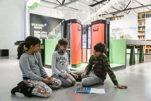 Drei Kinder sitzen in der Ausstellung auf dem Boden um ein aufgeschlagenes Heft herum. Im Hintergrund sieht man einen großen Computer mit rotem Gehäuse. Das gleiche Objekt erkennt man in dem Heft.