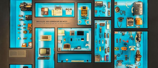 A wall-size display case composed of rectangular sections, some large and some small. Each section features communication devices from various ages. The back of the display case is bright blue.