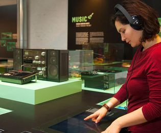 A woman wearing large headphones stands at a table that has display cases and objects on it. The word “music” can be seen on the wall in the background.