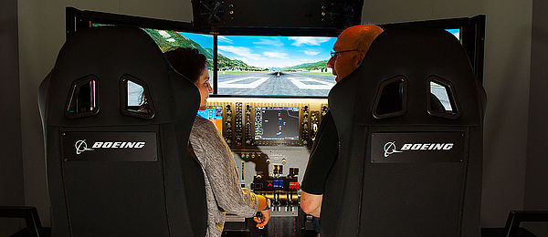 Blick von hinten in einen Flugsimulator: In dem rechten Sessel sitzt der Fluglehrer, im linken Sitz die Flugschülerin. Vor den beiden ist ein dreigeteilter Monitor zu sehen, auf dem man eine Rollbahn erkennen kann, außerdem ein Schaltboard und Steuerknüppel.